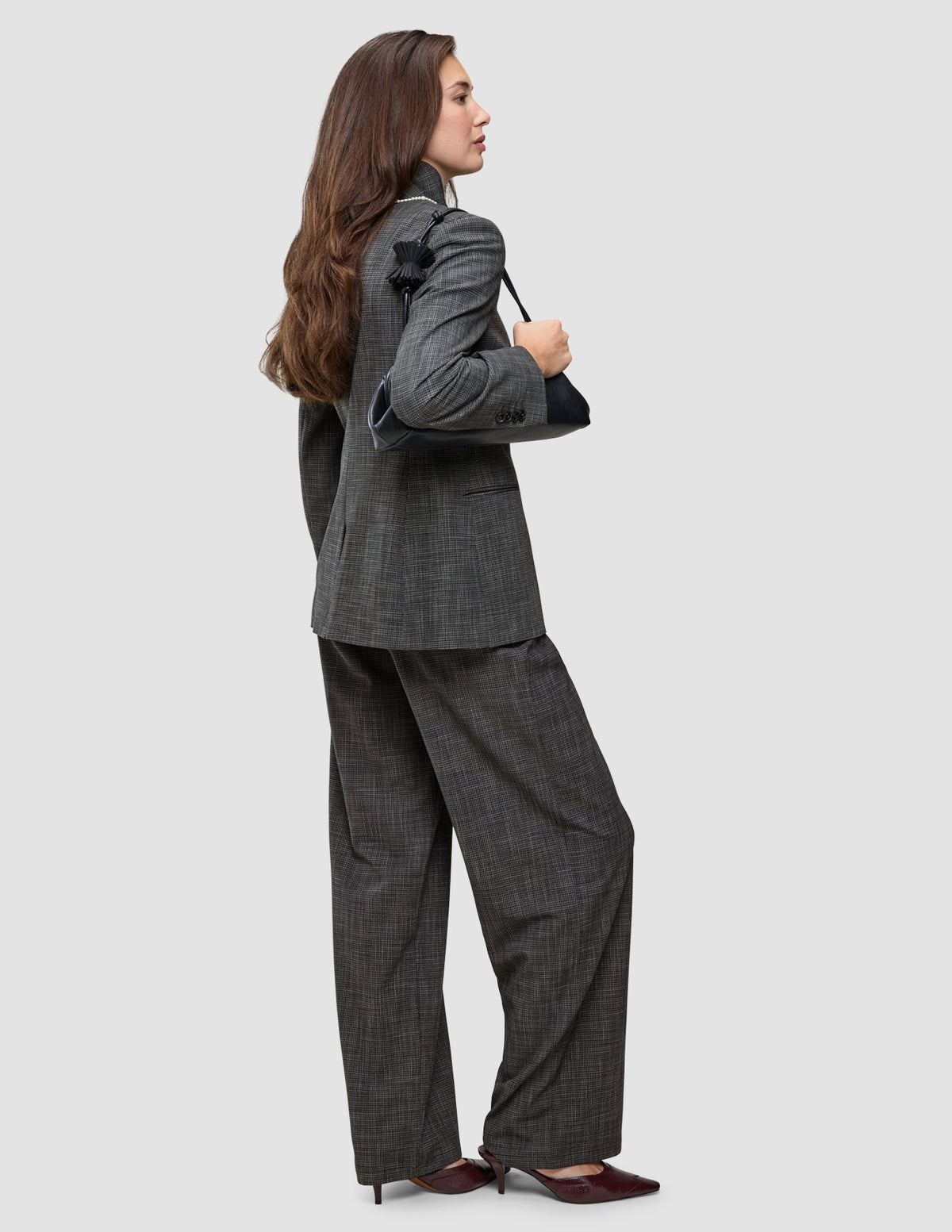 Eine Person, die einen Anzug mit auffälligem grauem Karomuster trägt, bestehend aus einem Blazer und einer dazu passenden Hose, und dabei eine schwarze Umhängetasche bei sich hat.