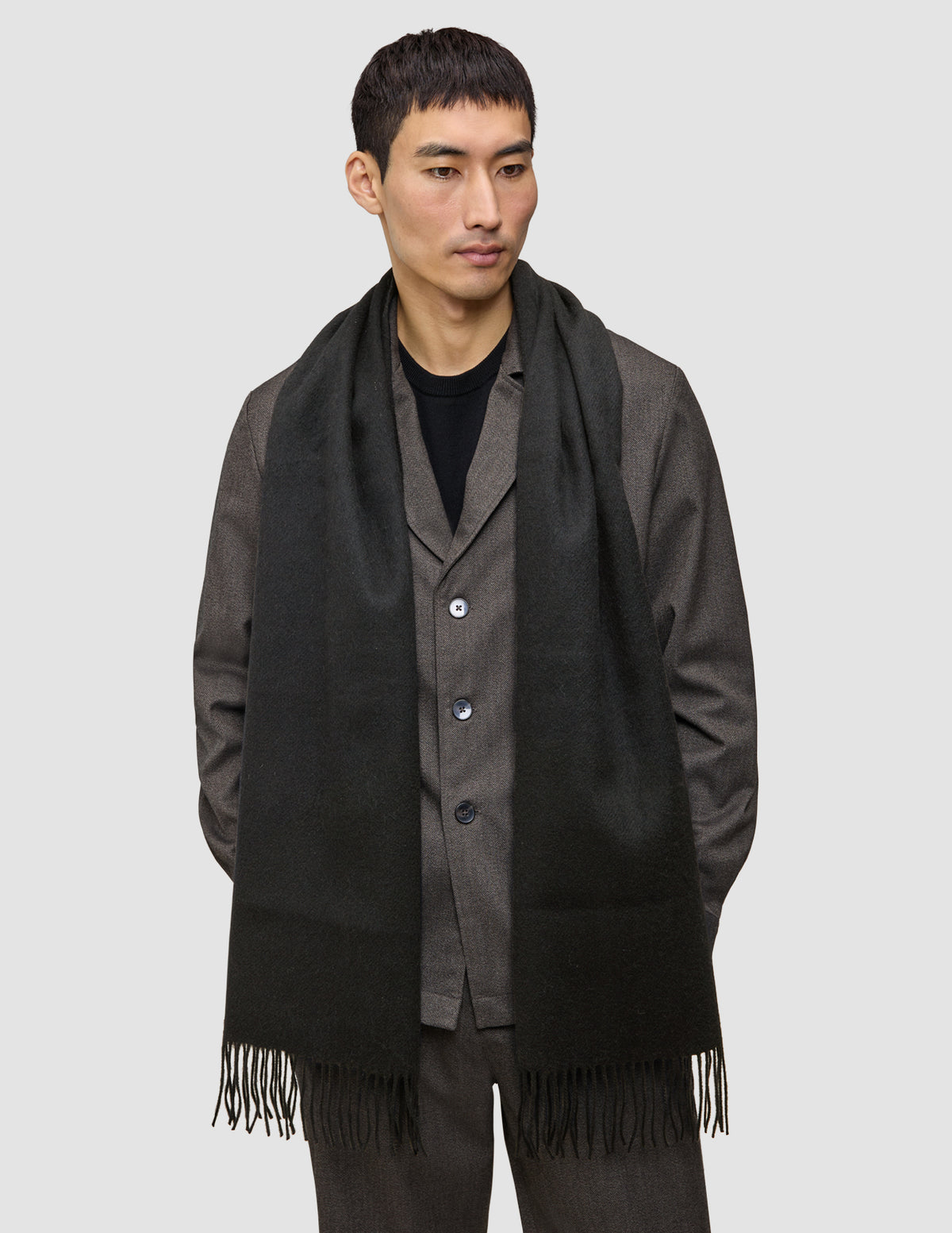 Ein Mann trägt eine dunkelbraune Jacke über einem schwarzen Hemd und einen Scarf Black, einen langen, mit Fransen verzierten Schal aus Kaschmirwolle im minimalistischen skandinavischen Design.