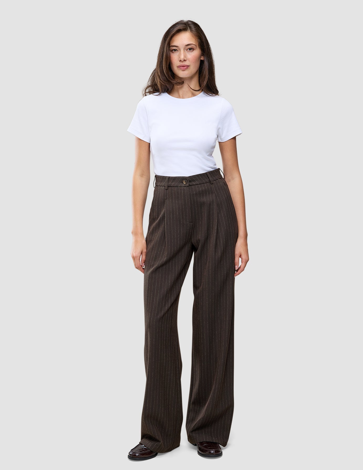 Eine Person, die ein weißes T-Shirt und eine weit geschnittene Serene Pants Wide Walnut Herringbone mit vertikalen Nadelstreifen trägt.