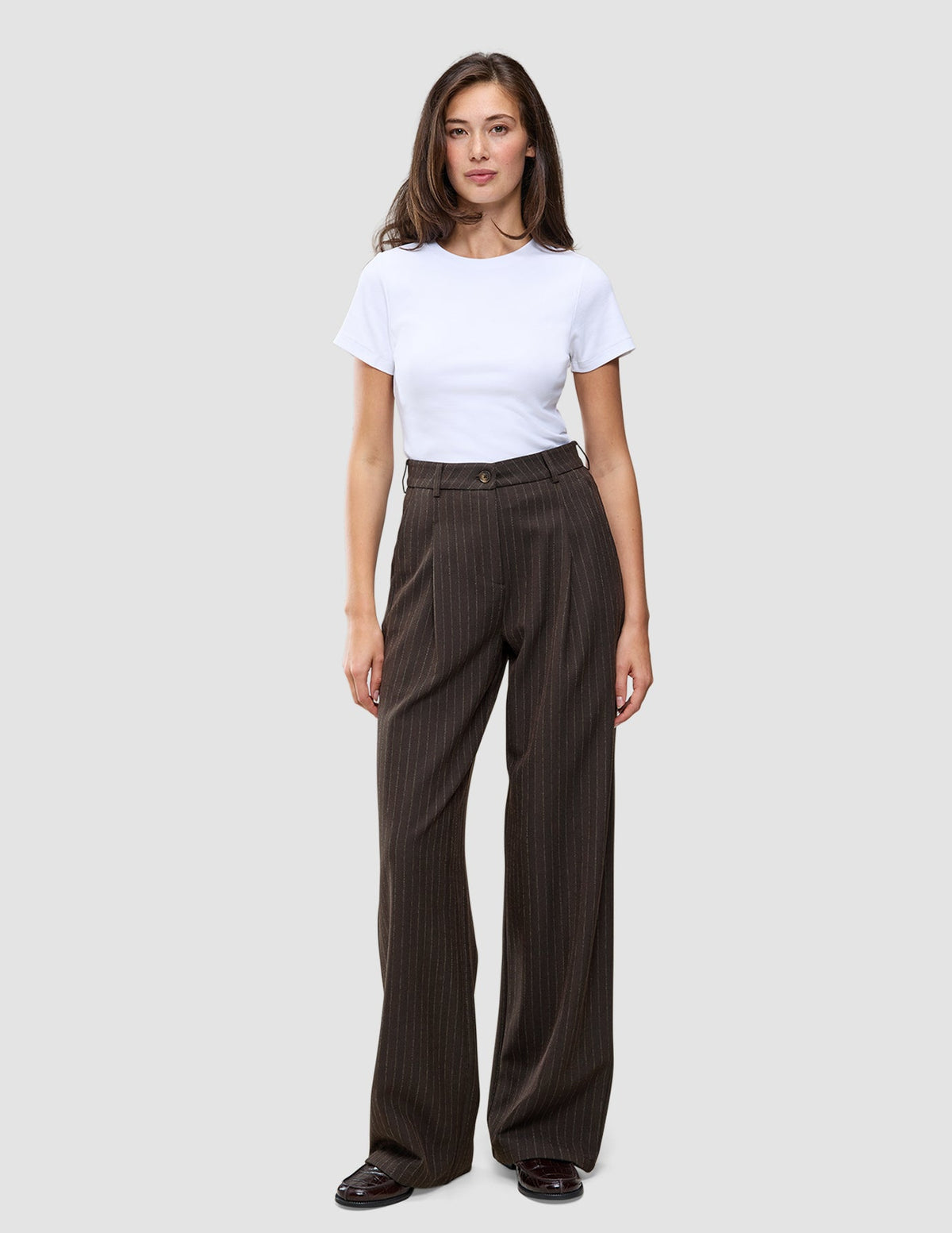 Eine Person, die ein weißes T-Shirt und eine weit geschnittene Serene Pants Wide Walnut Herringbone mit vertikalen Nadelstreifen trägt.
