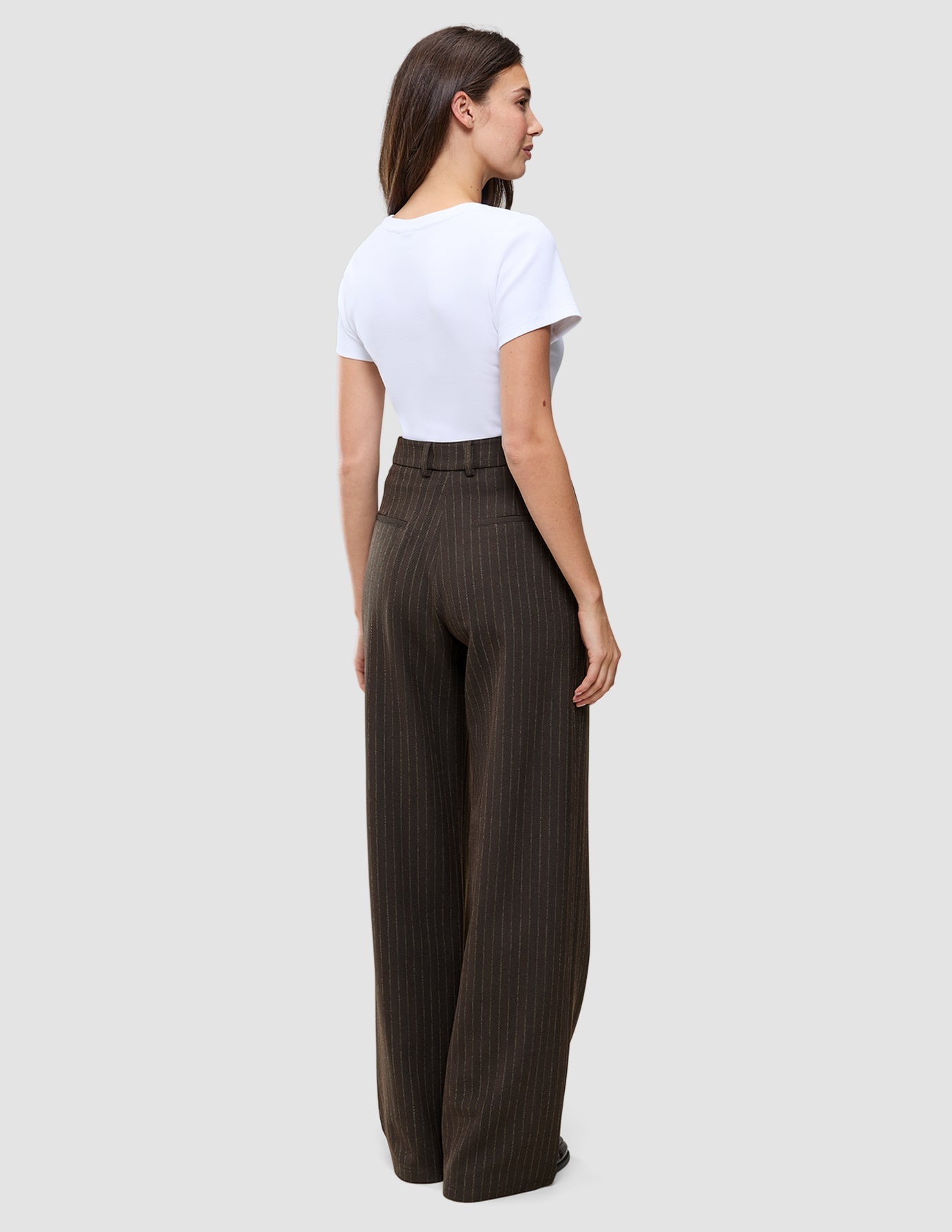 Eine Frau, von hinten gesehen, trägt ein weißes T-Shirt und eine braune Serene Pants Wide Walnut Herringbone mit weitem Bein und dezentem vertikalem Nadelstreifenmuster, die das minimalistische skandinavische Design verkörpert.