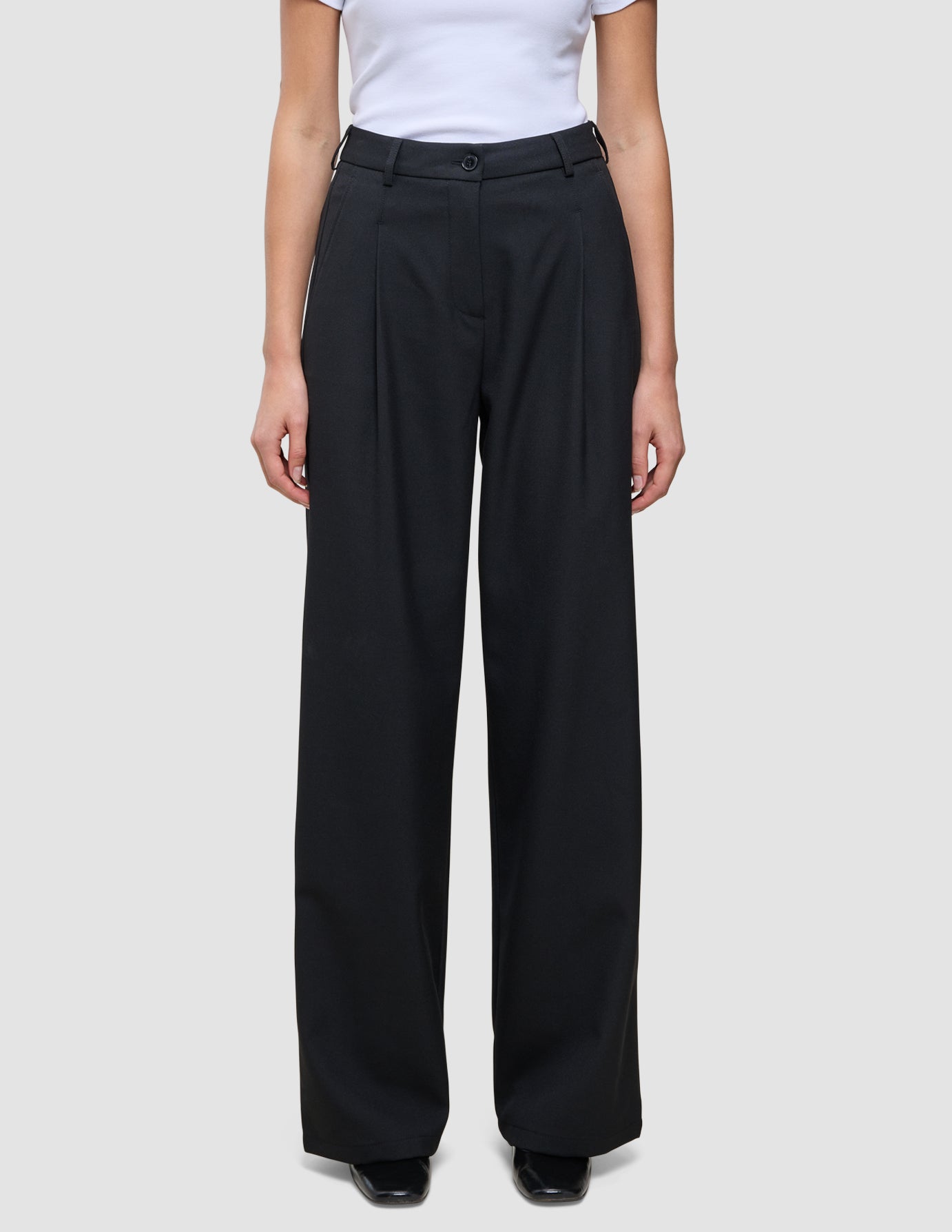 Eine Person, die eine hoch taillierte schwarze Serene Pants Wide Black mit weitem Bein und Falten auf der Vorderseite trägt, dazu ein weißes, in die Hose gestecktes T-Shirt.