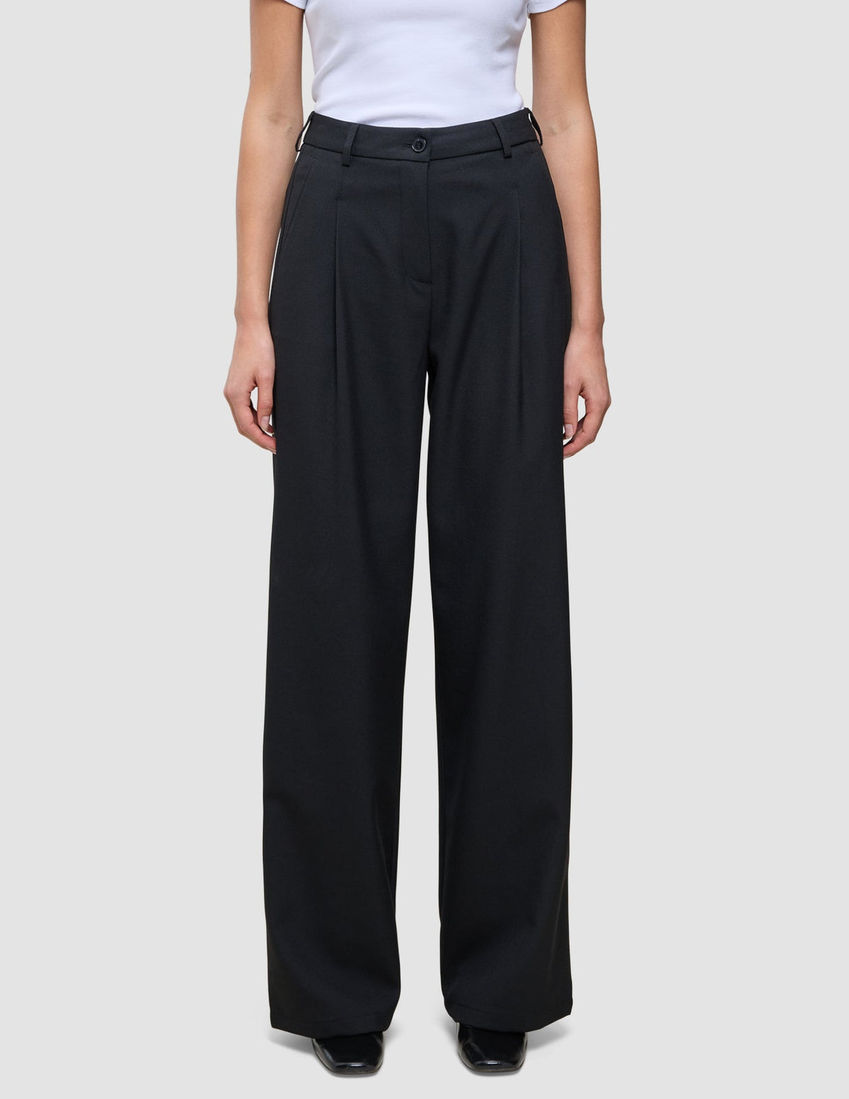 Eine Person, die eine hoch taillierte schwarze Serene Pants Wide Black mit weitem Bein und Falten auf der Vorderseite trägt, dazu ein weißes, in die Hose gestecktes T-Shirt.