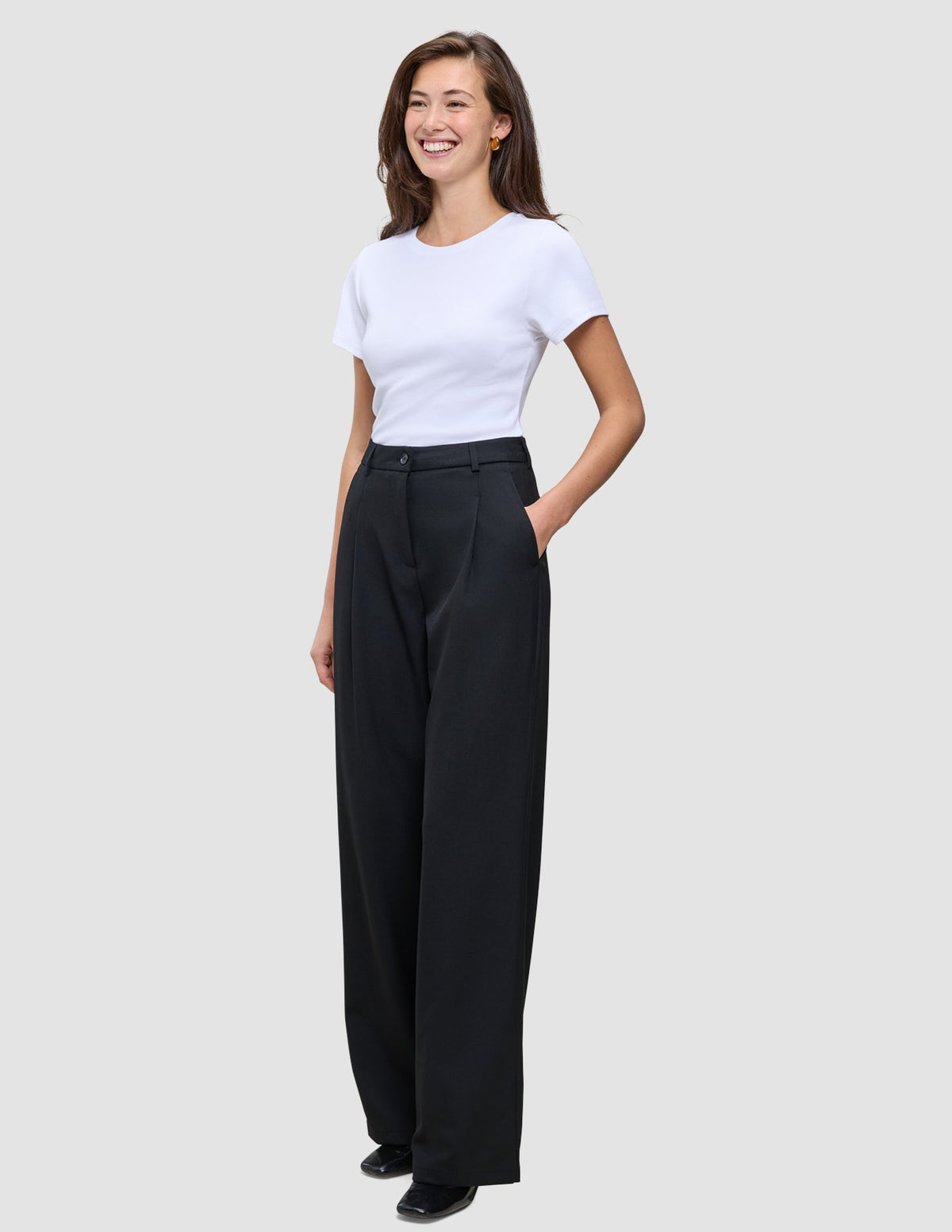 Eine Frau trägt ein weißes T-Shirt, das sie in eine hoch taillierte, weit geschnittene Serene Pants Wide Black gesteckt hat.