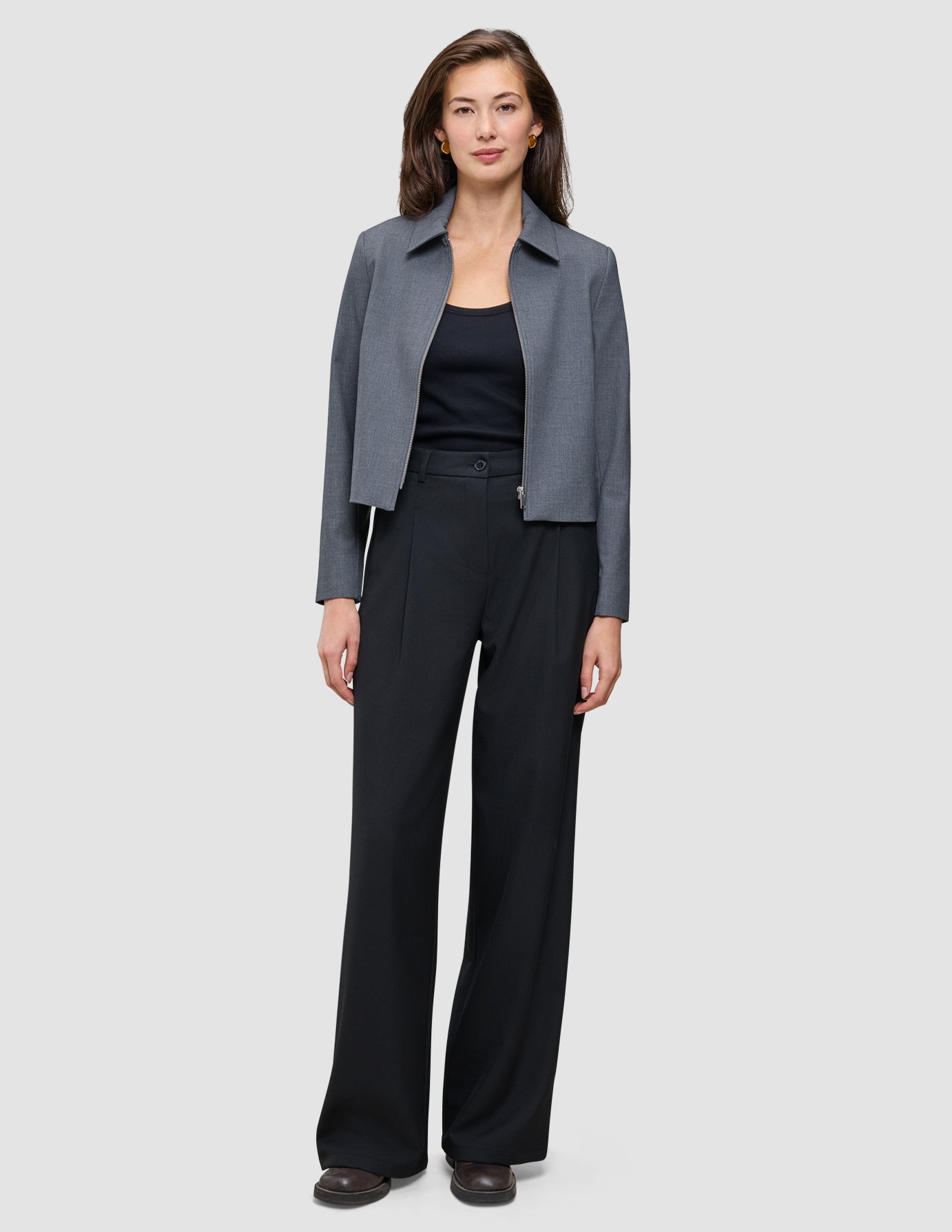 Eine Frau, die eine graue Jacke mit Reißverschluss, ein schwarzes Trägertop und eine schwarze Hose mit weitem Bein vom Modell Serene Pants Wide Black trägt.