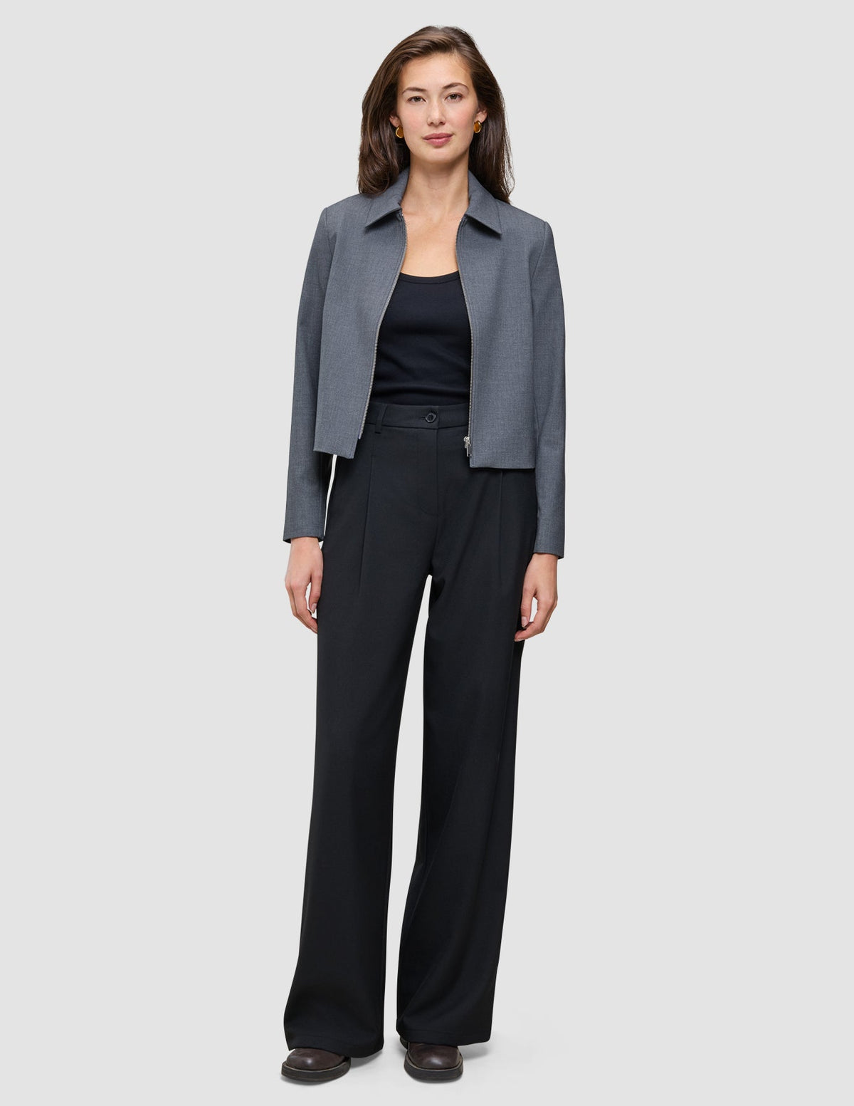 Eine Frau, die eine graue Jacke mit Reißverschluss, ein schwarzes Trägertop und eine schwarze Hose mit weitem Bein vom Modell Serene Pants Wide Black trägt.