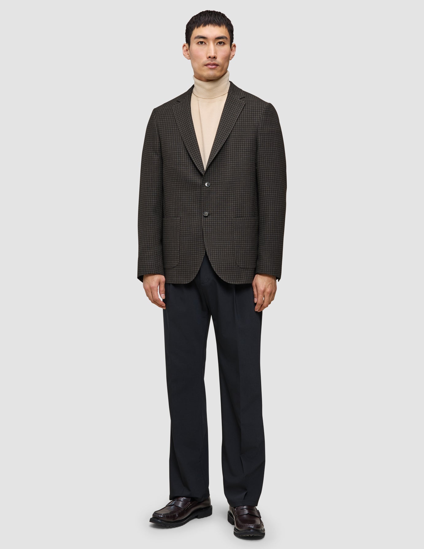 Ein Mann im Strick-und-Blazer-Look mit einem Wollblazer über einem beigen Merino-Strickpullover, einer dunklen Hose und dunklen Leder-Loafern.