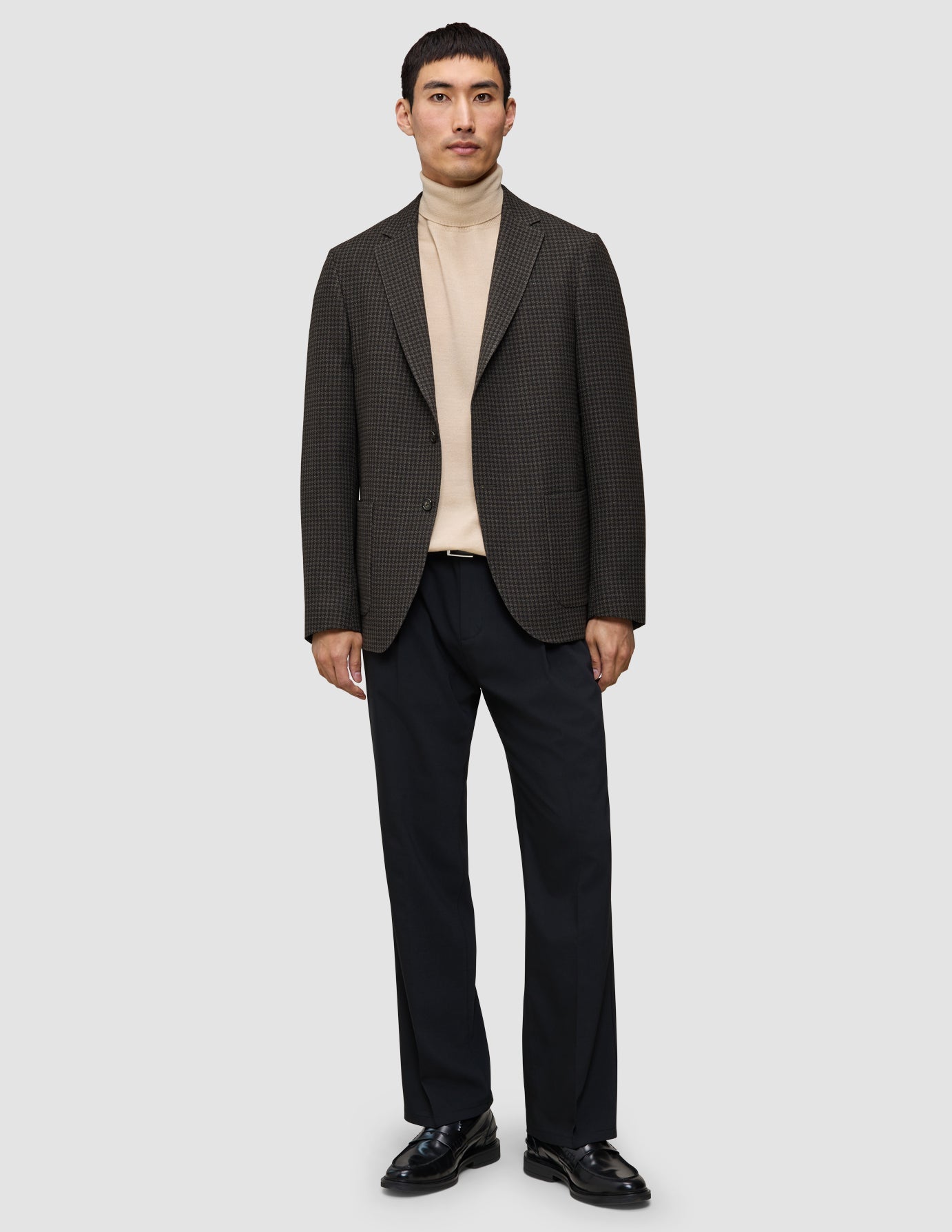 Ein Mann im Strick-und-Blazer-Look: ein hellbrauner Merino-Strickpullover, ein zeitloser Wollblazer, eine dunkle Hose und schwarze Loafer.