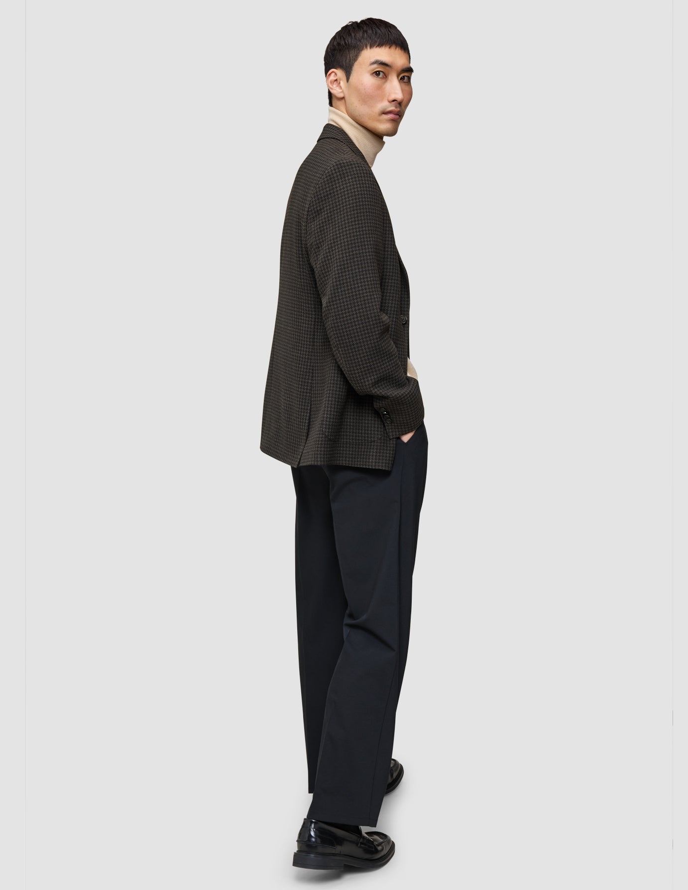 Ein Mann im Strick-und-Blazer-Look – bestehend aus einem Merino-Strickpullover, einem dunkel gemusterten Blazer und einer dunklen Hose – blickt über seine Schulter.