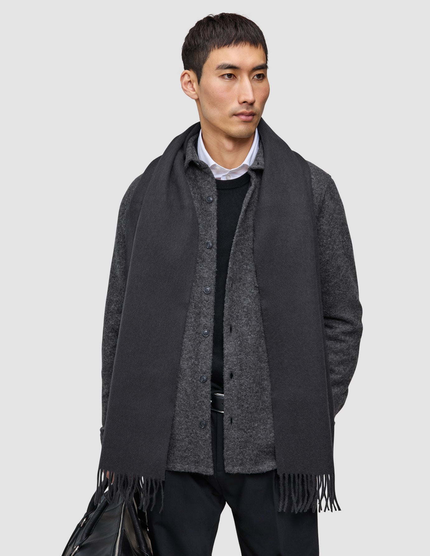 Ein Mann, der ein gelungenes Lagen-Outfit trägt: einen grauen Premium-Merino-Strickschal über einem dunkelgrauen doppellagigen Woll-Overshirt und einem schwarzen Hemd; er hält eine schwarze Tasche in der Hand.
