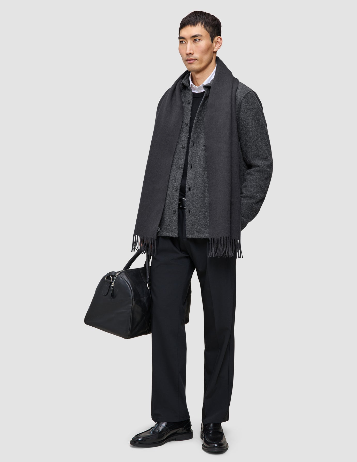 Ein Mann, der ein gelungeneres Schichten-Outfit trägt, bestehend aus einem doppellagigen Wollhemd über einem hochwertigen Merino-Pullover, einem schwarzen Schal, einer Hose und Lederschuhen, der eine schwarze Reisetasche in der Hand hält.