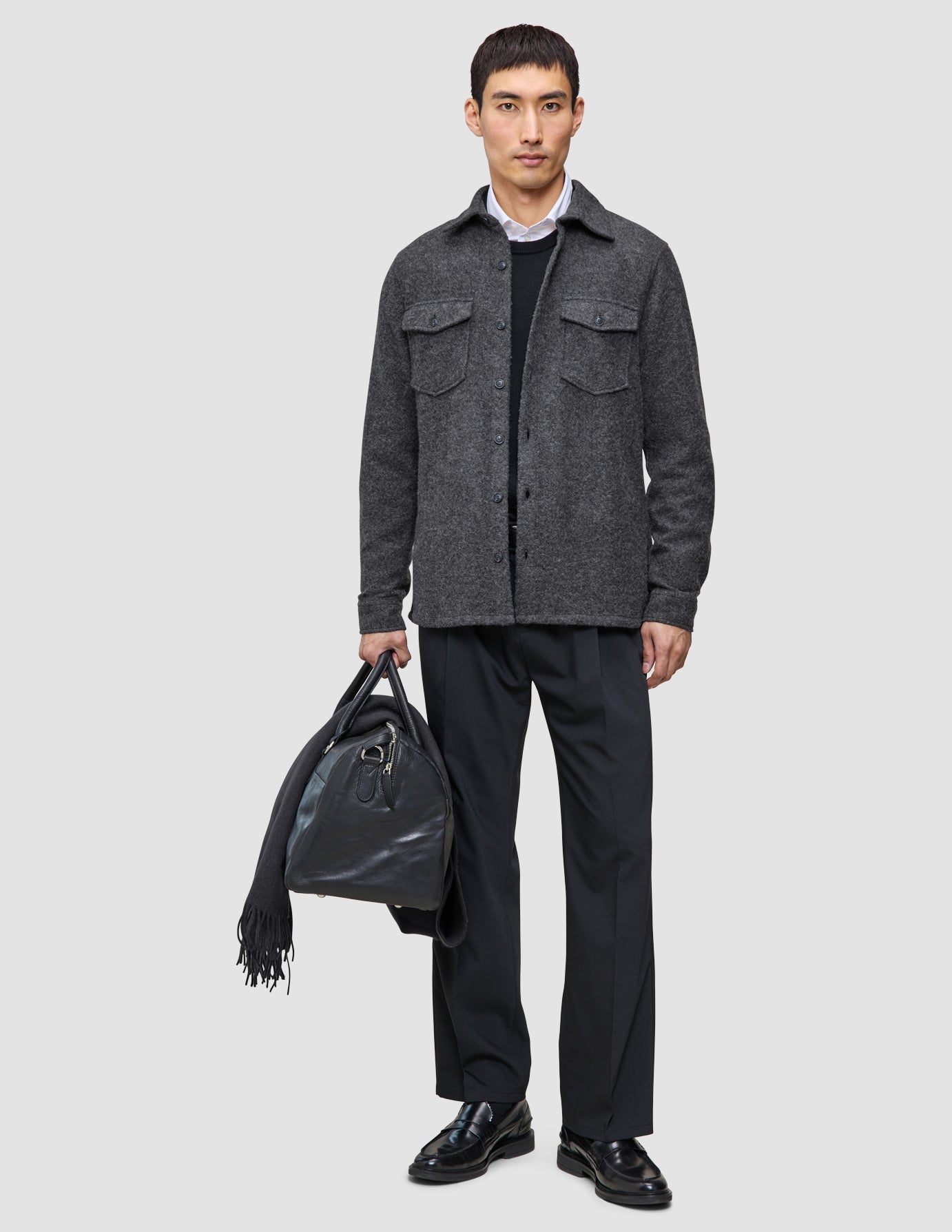 Ein Mann, der ein graues Layers that work-Überhemd, ein schwarzes Hemd und eine Tech Wool-Hose trägt und eine schwarze Leder-Reisetasche hält, über deren Griff ein Schal drapiert ist.