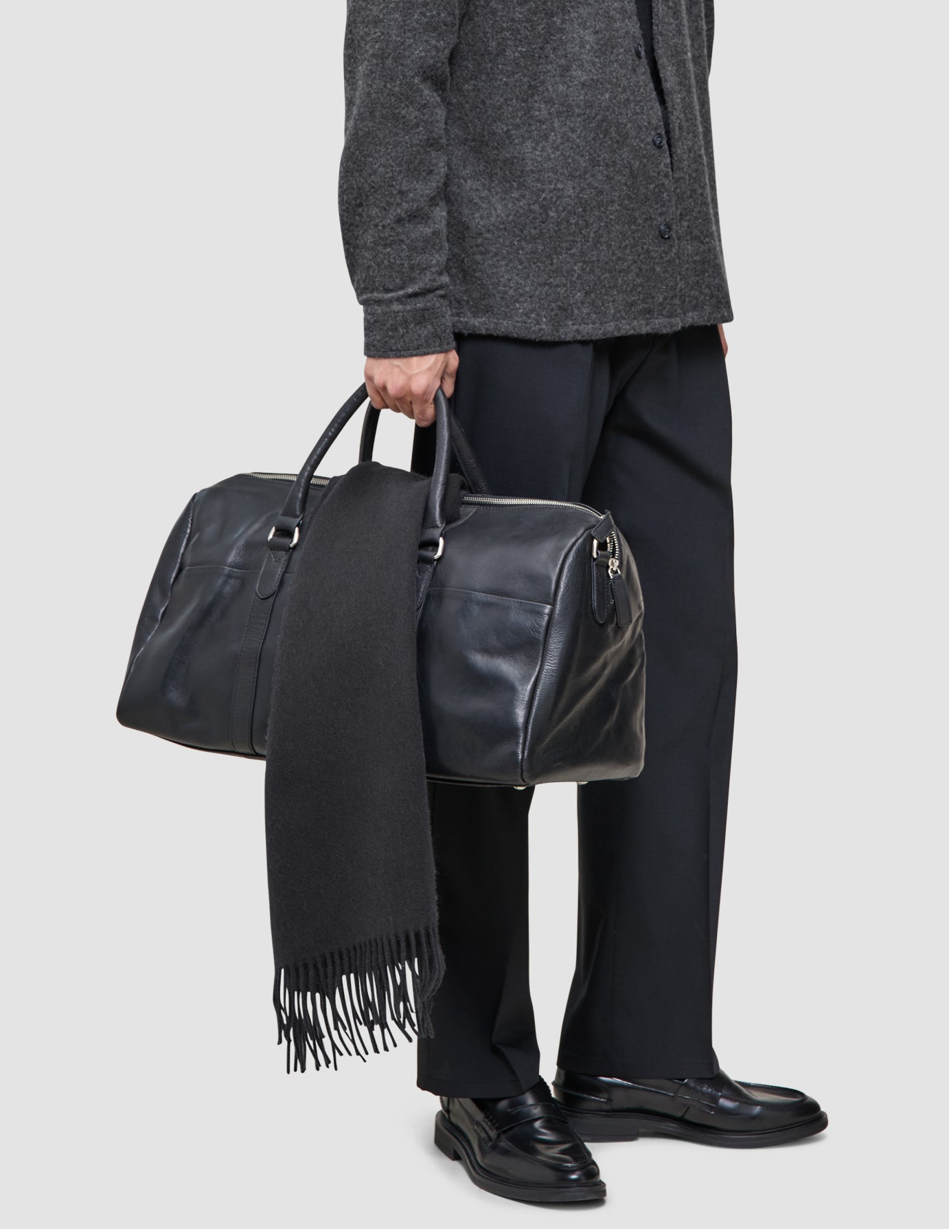 Eine Person, die ein gut abgestimmtes Lagen-Outfit trägt – bestehend aus einem grauen Premium-Merino-Strickpullover und einer schwarzen Tech-Wool-Hose –, hält eine schwarze Leder-Reisetasche in der Hand, über deren Griff ein schwarzer Schal drapiert ist.