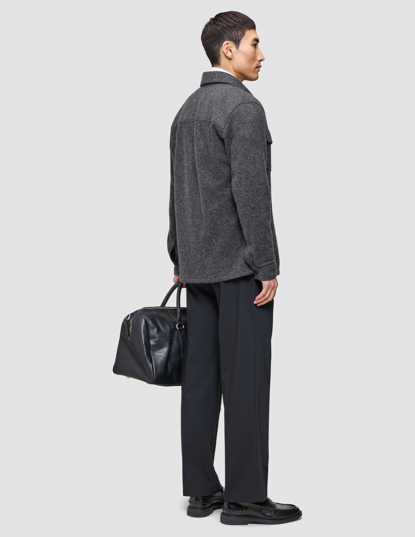 Ein Mann trägt ein gelungenes Mehrschicht-Outfit, bestehend aus einem grauen, doppellagigen Woll-Overshirt über einem Hemd mit Kragen und einer schwarzen Tech-Wollhose, und hält dabei eine schwarze Leder-Reisetasche in der Hand.