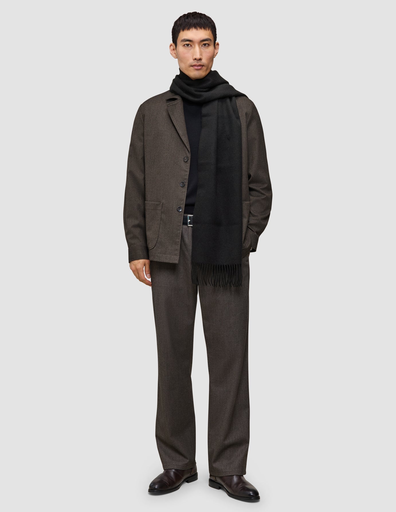 Ein Mann, der eine etwas andere Art von Anzug trägt, bestehend aus einem Overshirt und einer Hose von Atelier, die er über einem hochwertigen Merino-Strickpullover und einem Schal trägt.