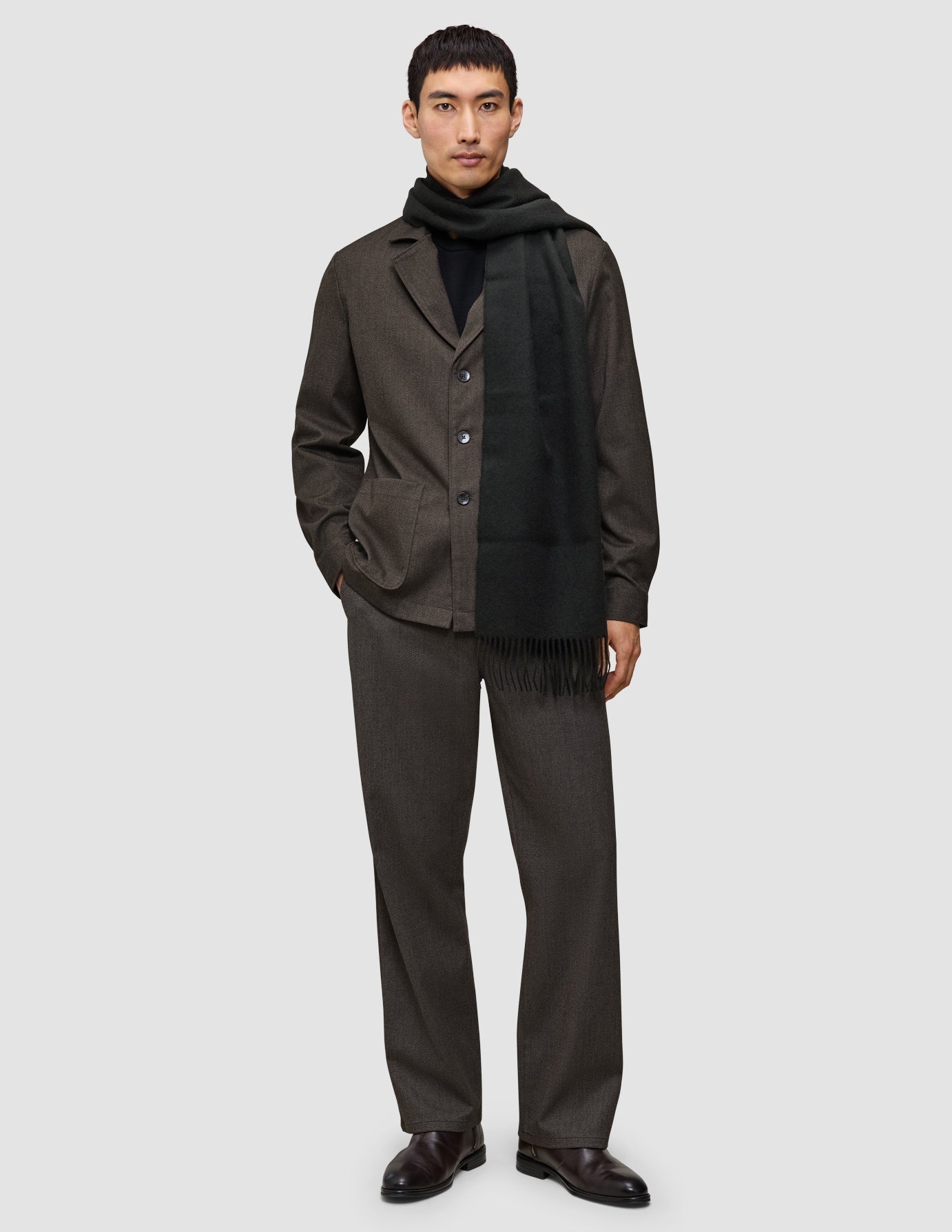 Ein Mann, der eine etwas andere Art von Anzug trägt: ein dunkelbraunes Atelier Overshirt und eine dazu passende Atelier Pants über einem schwarzen Rollkragenpullover, ergänzt durch einen schwarzen Schal und dunkle Lederschuhe.