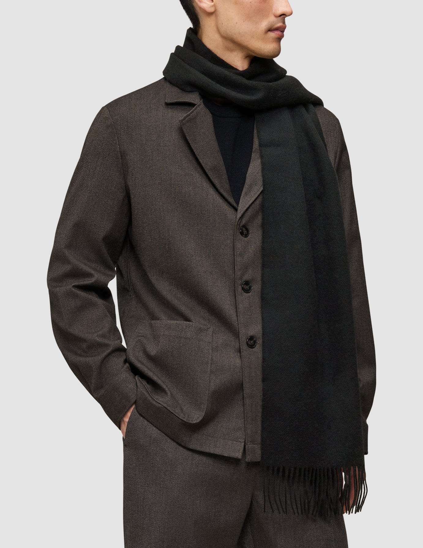 Ein Mann, der einen braunen, etwas andersartigen Anzug über einem schwarzen Premium-Merino-Strickpullover und einem schwarzen Schal mit Fransen trägt.