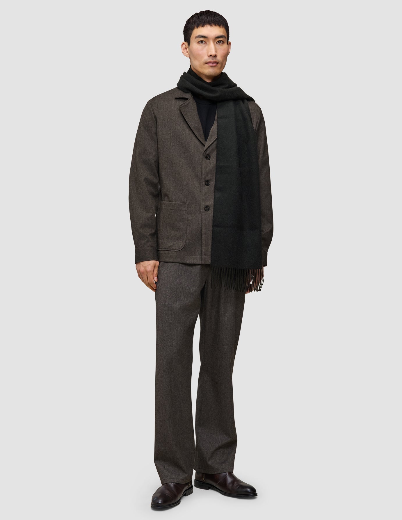 Eine Person, die einen etwas anderen Anzug in Dunkelbraun trägt, bestehend aus einem Atelier Overshirt und einer dazu passenden Atelier Pants, kombiniert mit einem schwarzen Rollkragenpullover und einem Schal.