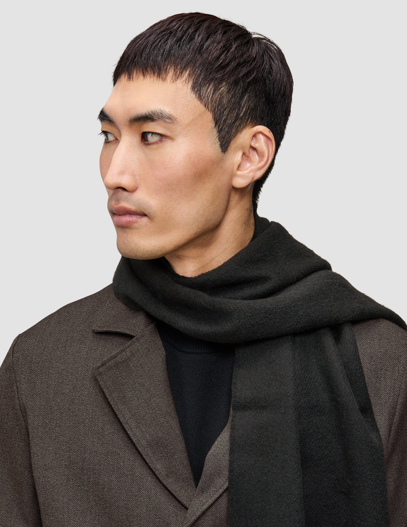 Ein Mann, der ein A different kind of suit Overshirt von Atelier über einem Premium-Merino-Strickpullover trägt und einen dunkelgrünen Schal um den Hals gelegt hat.