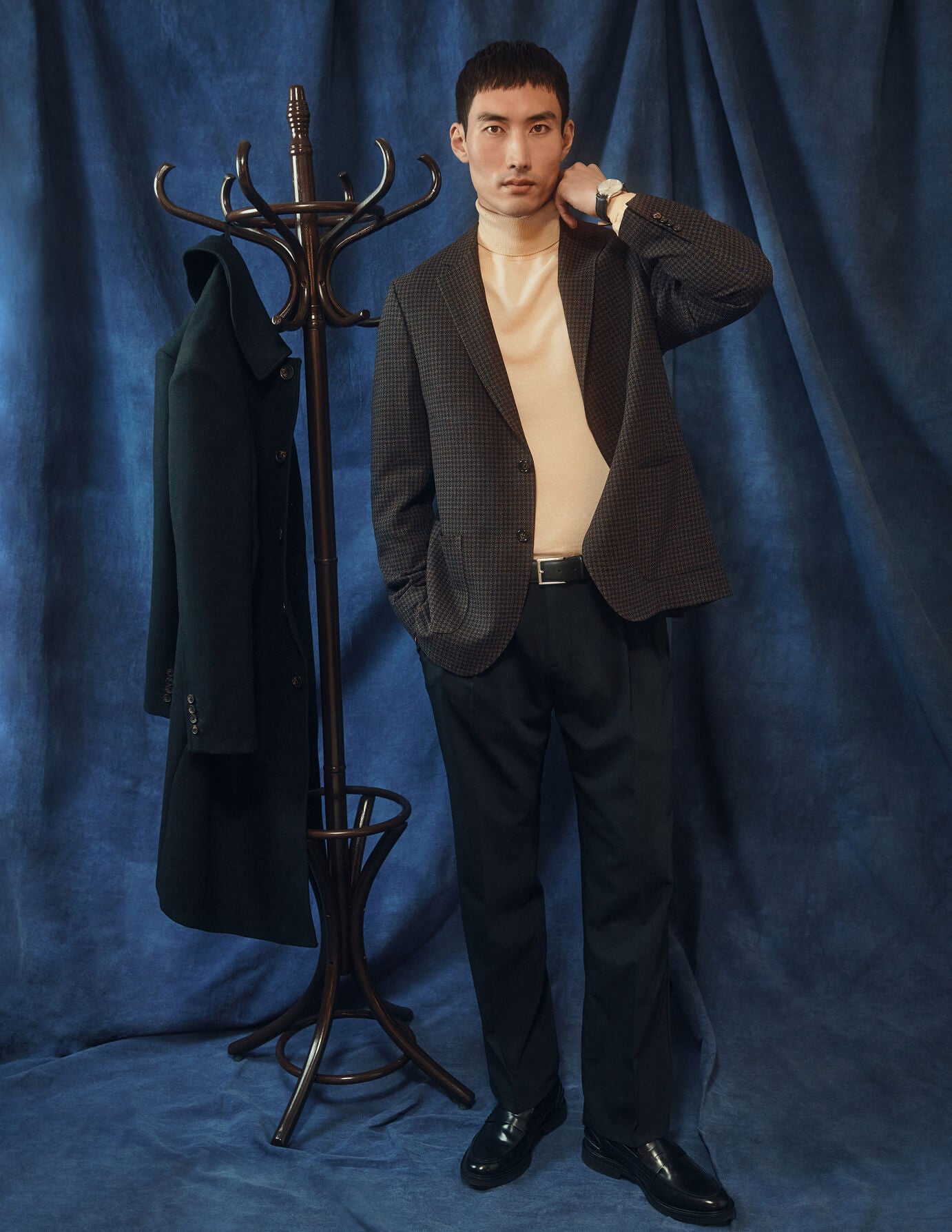Ein Mann, der einen gemusterten Blazer, einen cremefarbenen Rollkragenpullover und eine schwarze Tech-Wool-Anzughose in Relaxed Fit trägt, steht neben einer Garderobe, auf der ein dunkler Mantel hängt.