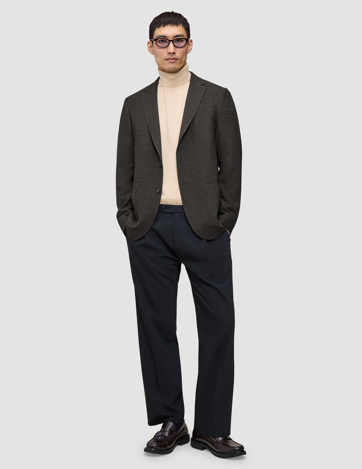 Eine Person, die einen Timeless Wool Blazer Slim in dunkelbraunem Karomuster, einen beigen Rollkragenpullover, eine schwarze Hose und Loafer trägt.