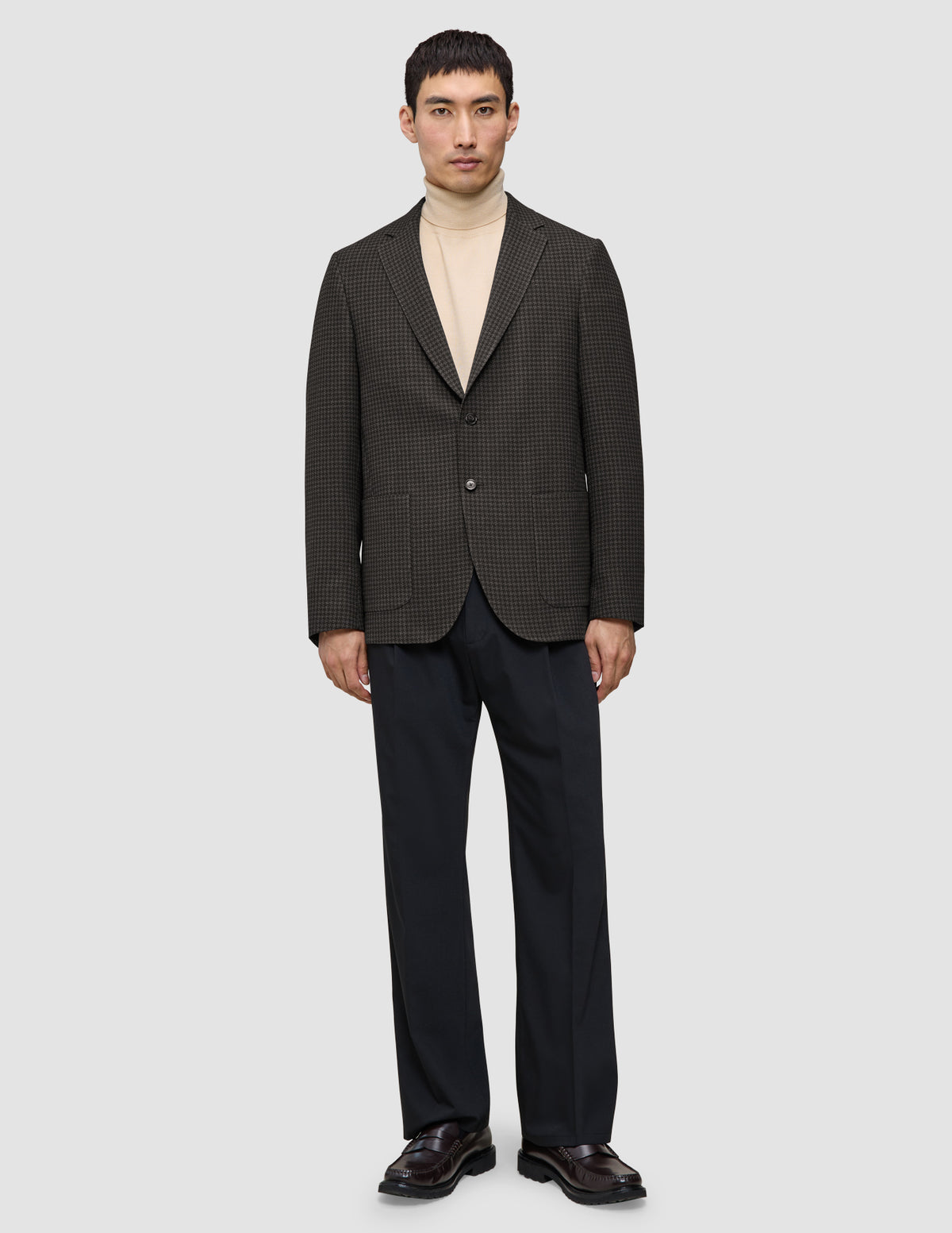 Ein Mann trägt einen dunkelbraunen, karierten Timeless Wool Blazer Slim über einem beigen Pullover mit Stehkragen und einer dunklen Hose.