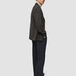 Ein Mann, der einen dunkelbraunen, karierten Timeless Wool Blazer Slim über einem beigen Rollkragenpullover und einer dunklen Hose trägt, blickt über seine Schulter.