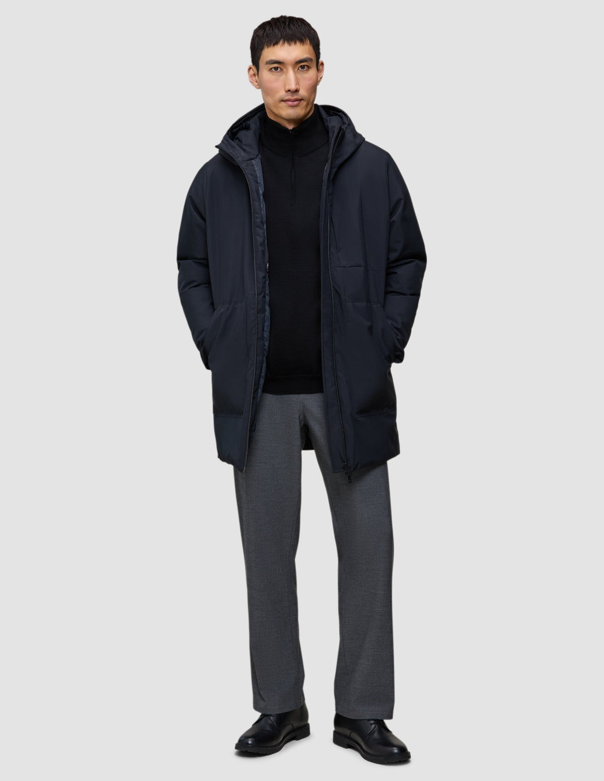 Ein Mann, der eine schwarze Utility-Jacke aus recyceltem Polyester über einem schwarzen Pullover mit Reißverschluss am Kragen und einer grauen Hose trägt.