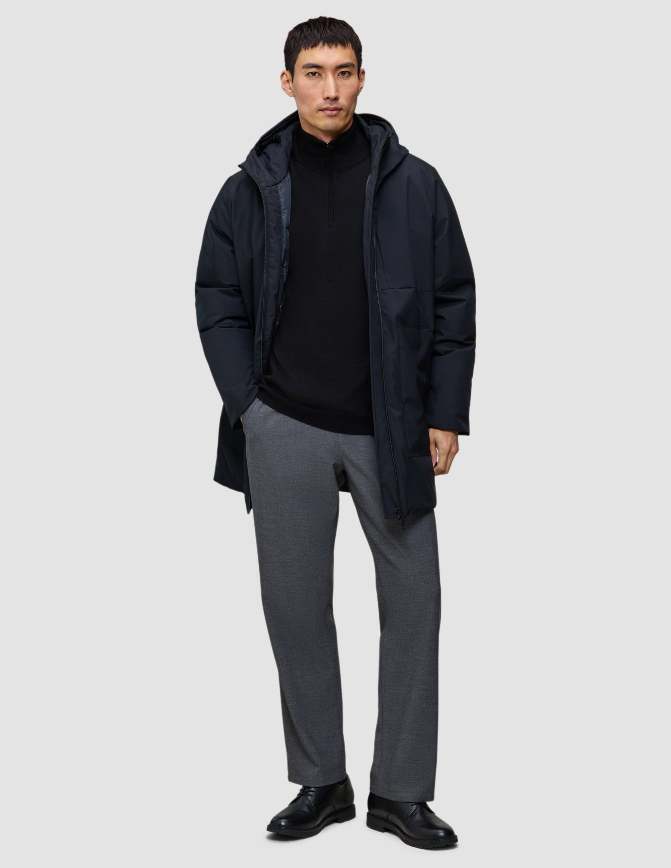 Eine Person, die eine schwarze Utility-Jacke mit Kapuze aus recyceltem Polyester über einem schwarzen Pullover mit Viertelreißverschluss, einer grauen Hose und schwarzen Anzugschuhen trägt.