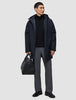 Ein Mann, der eine schwarze Utility-Jacke über einem schwarzen Pullover mit Viertelreißverschluss und einer grauen Hose trägt und eine schwarze Ledertasche in der Hand hält.