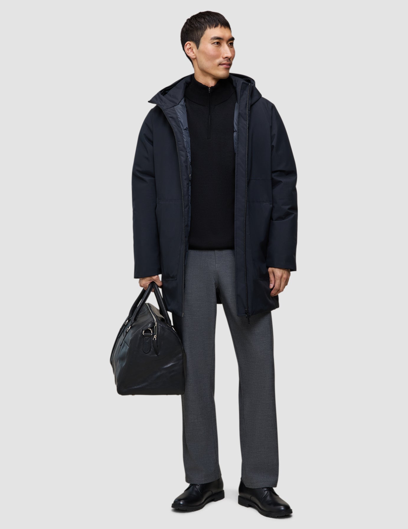 Ein Mann, der eine schwarze Utility-Jacke über einem schwarzen Pullover mit Viertelreißverschluss und einer grauen Hose trägt und eine schwarze Ledertasche in der Hand hält.