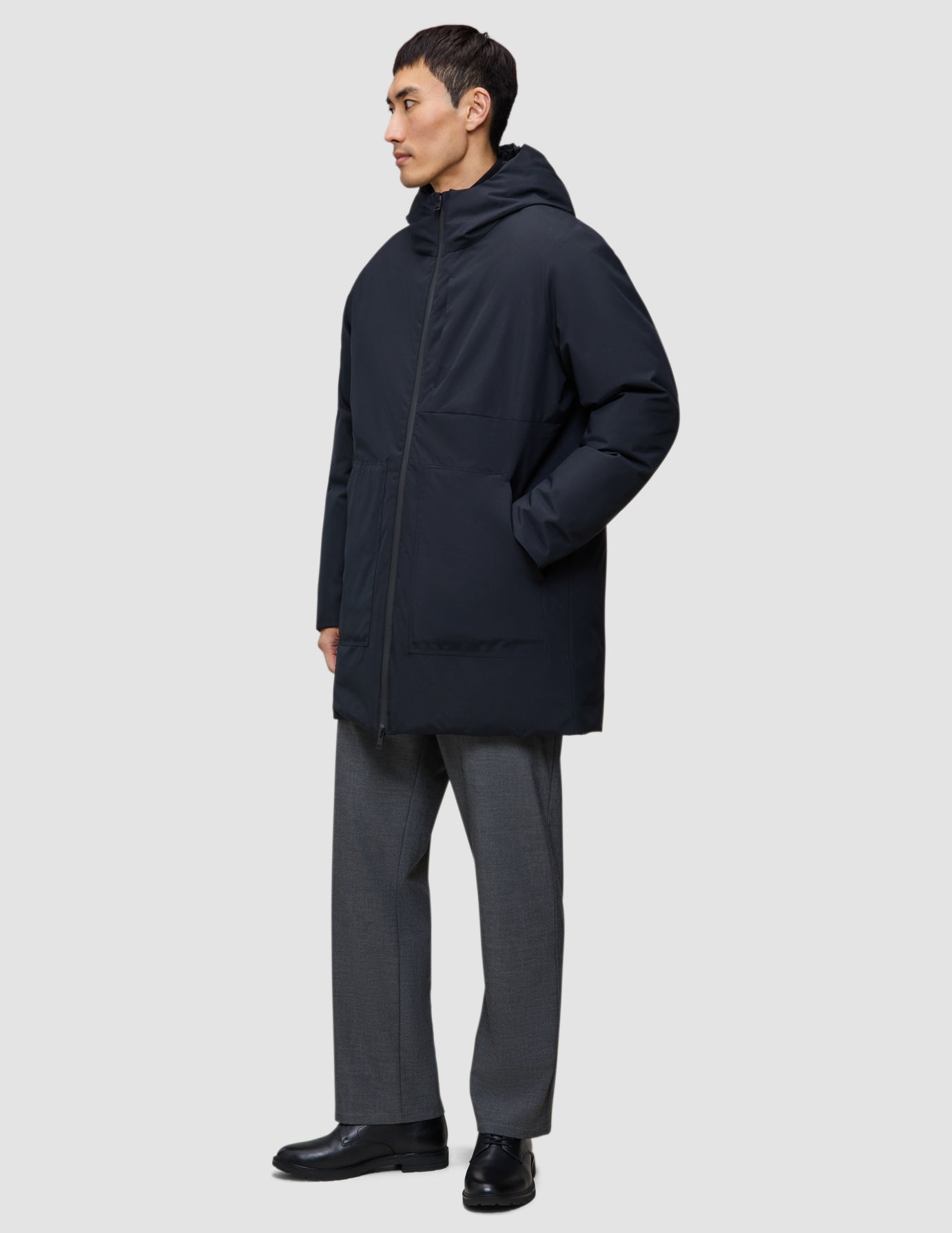 Eine Person, die eine schwarze Utility-Jacke mit Kapuze aus recyceltem Polyester und eine graue Hose trägt.