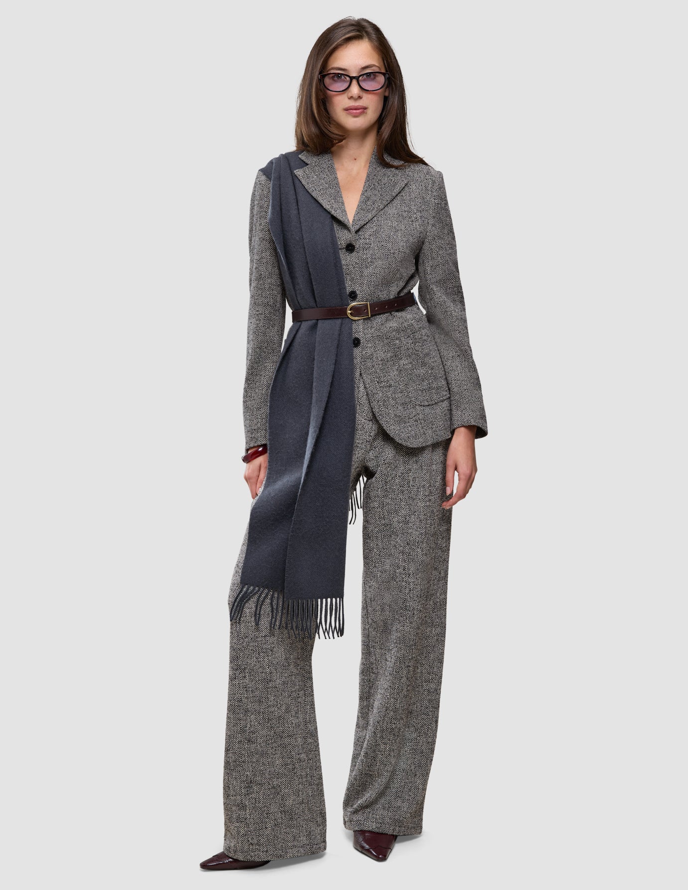 Eine Person trägt einen grauen Blazer aus Bouclé mit Fischgrätenmuster und dazu passende Women Ashryn Pants Wide Domino Herringbone, die mit einem dunkelbraunen Gürtel tailliert und durch einen langen, anthrazitfarbenen Schal mit Fransen akzentuiert werden.