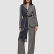 Eine Person trägt einen grauen Blazer aus Bouclé mit Fischgrätenmuster und dazu passende Women Ashryn Pants Wide Domino Herringbone, die mit einem dunkelbraunen Gürtel tailliert und durch einen langen, anthrazitfarbenen Schal mit Fransen akzentuiert werden.