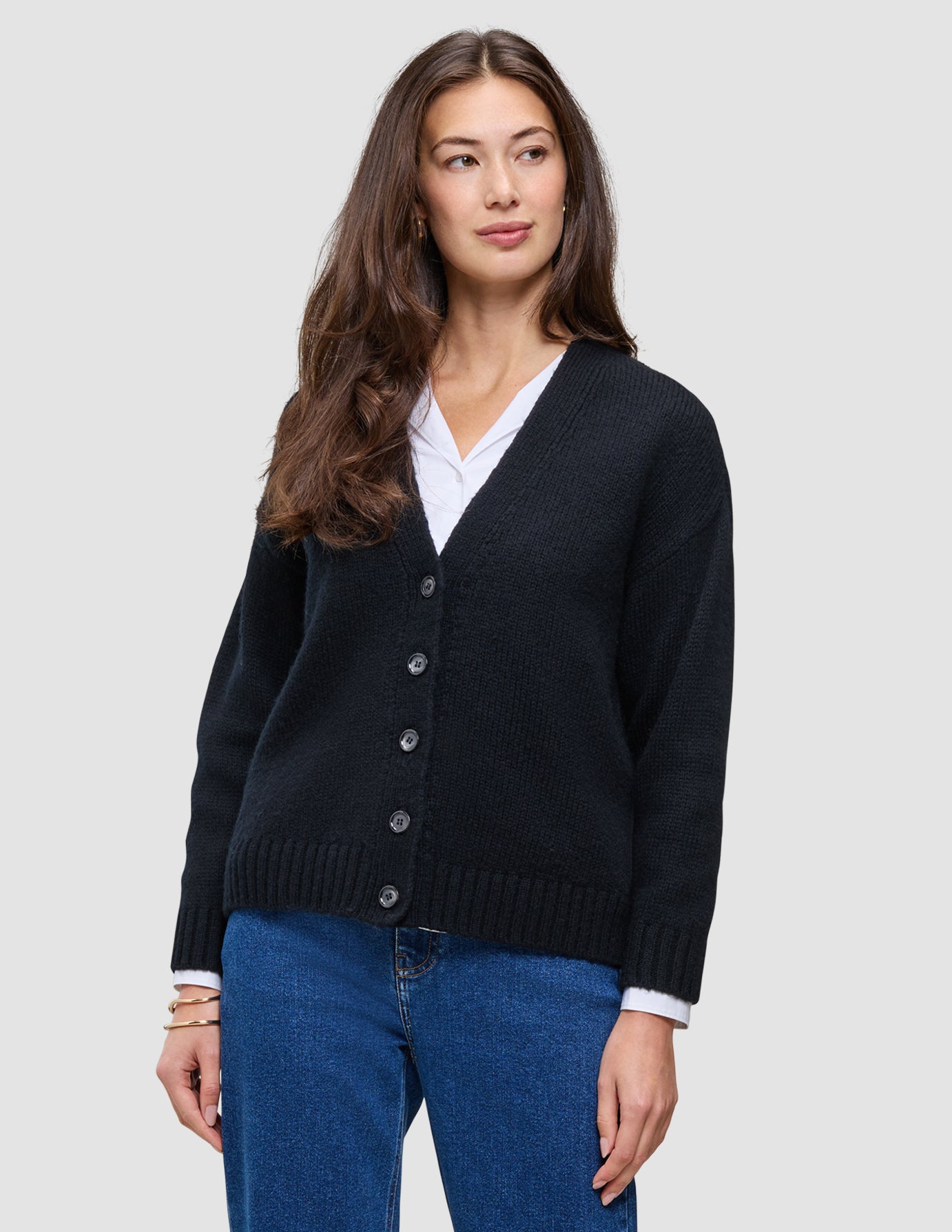 Eine Frau, die eine schwarze, grob gestrickte, weiche Wollstrickjacke für Damen über einem weißen Hemd mit Kragen und einer blauen Jeans trägt.