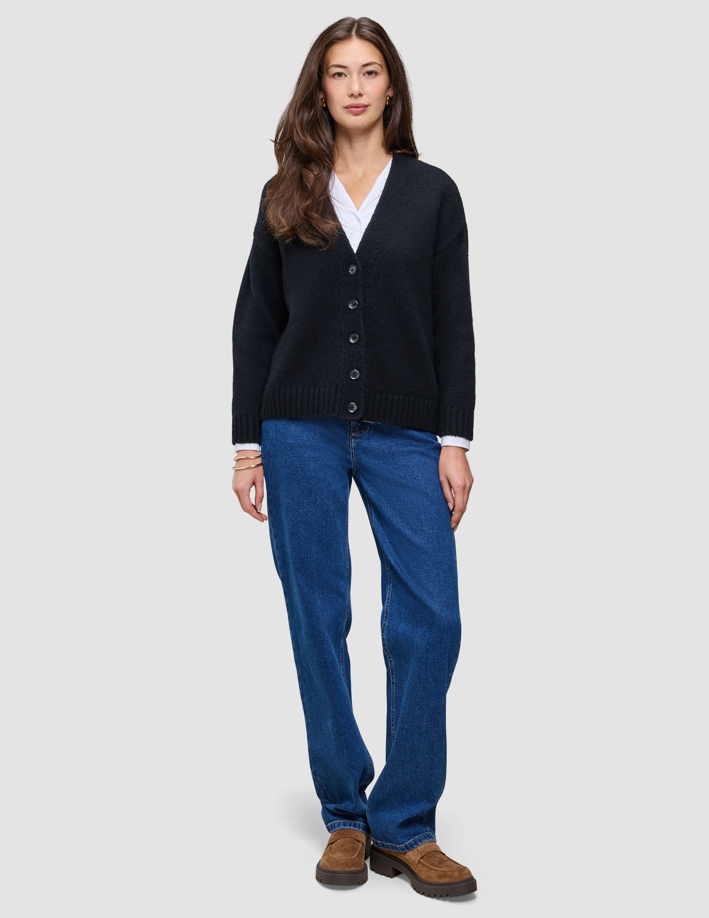 Eine Frau trägt eine schwarze, grob gestrickte, weiche Wollstrickjacke für Damen über einem weißen Hemd, dazu blaue Jeans und braune Slipper.