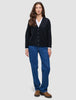 Eine Frau trägt eine schwarze, grob gestrickte, weiche Wollstrickjacke für Damen über einem weißen Hemd, dazu blaue Jeans und braune Slipper.