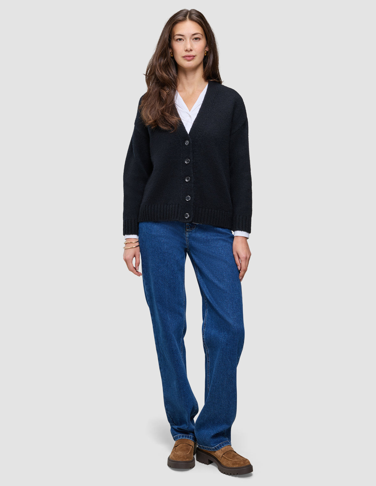 Eine Frau trägt eine schwarze, grob gestrickte, weiche Wollstrickjacke für Damen über einem weißen Hemd, dazu blaue Jeans und braune Slipper.