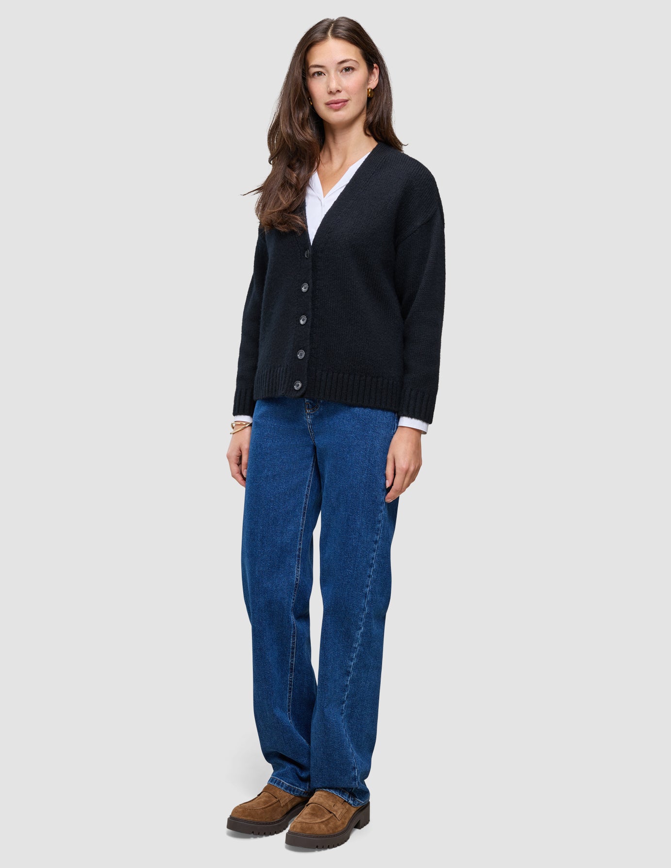 Eine Frau, die eine schwarze, grob gestrickte, weiche Wollstrickjacke für Damen über einem weißen Hemd, einer blauen Jeans und braunen Loafern trägt.