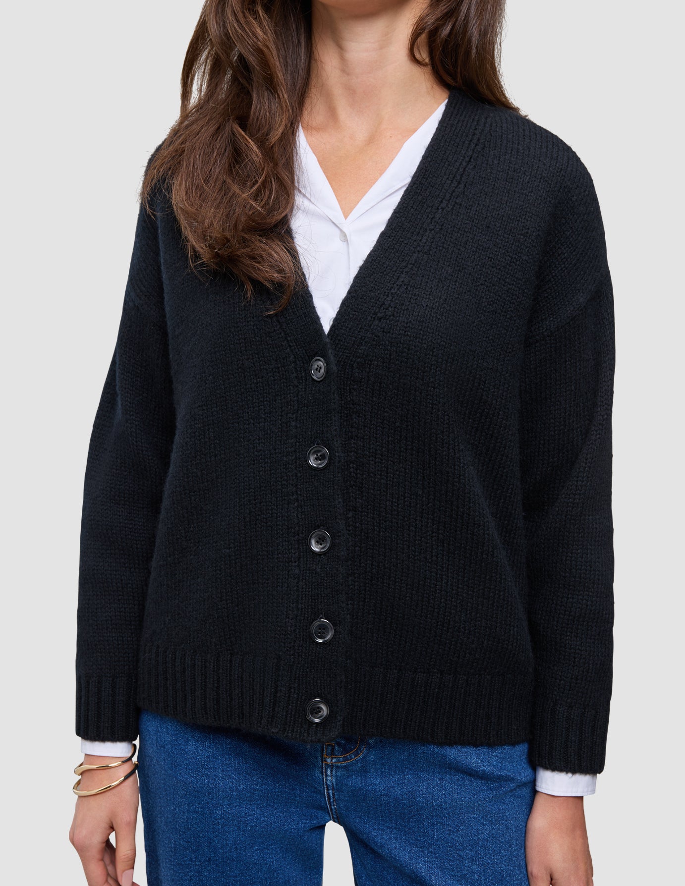 Eine Person, die eine schwarze, grob gestrickte, weiche Wollstrickjacke für Damen über einem weißen Hemd mit Kragen und einer blauen Jeans trägt.