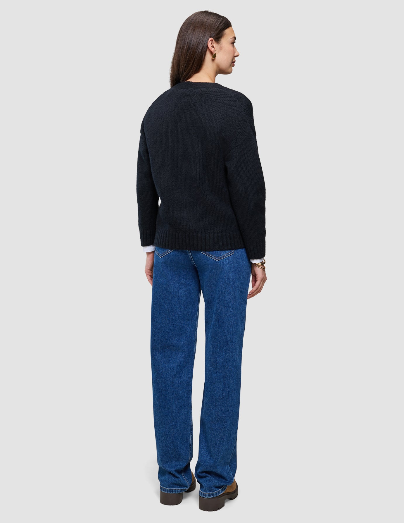 Eine Person, von hinten gesehen, die eine schwarze, dick gestrickte, weiche Damen-Strickjacke aus Wolle, ein weißes Hemd und eine blaue Jeans trägt.