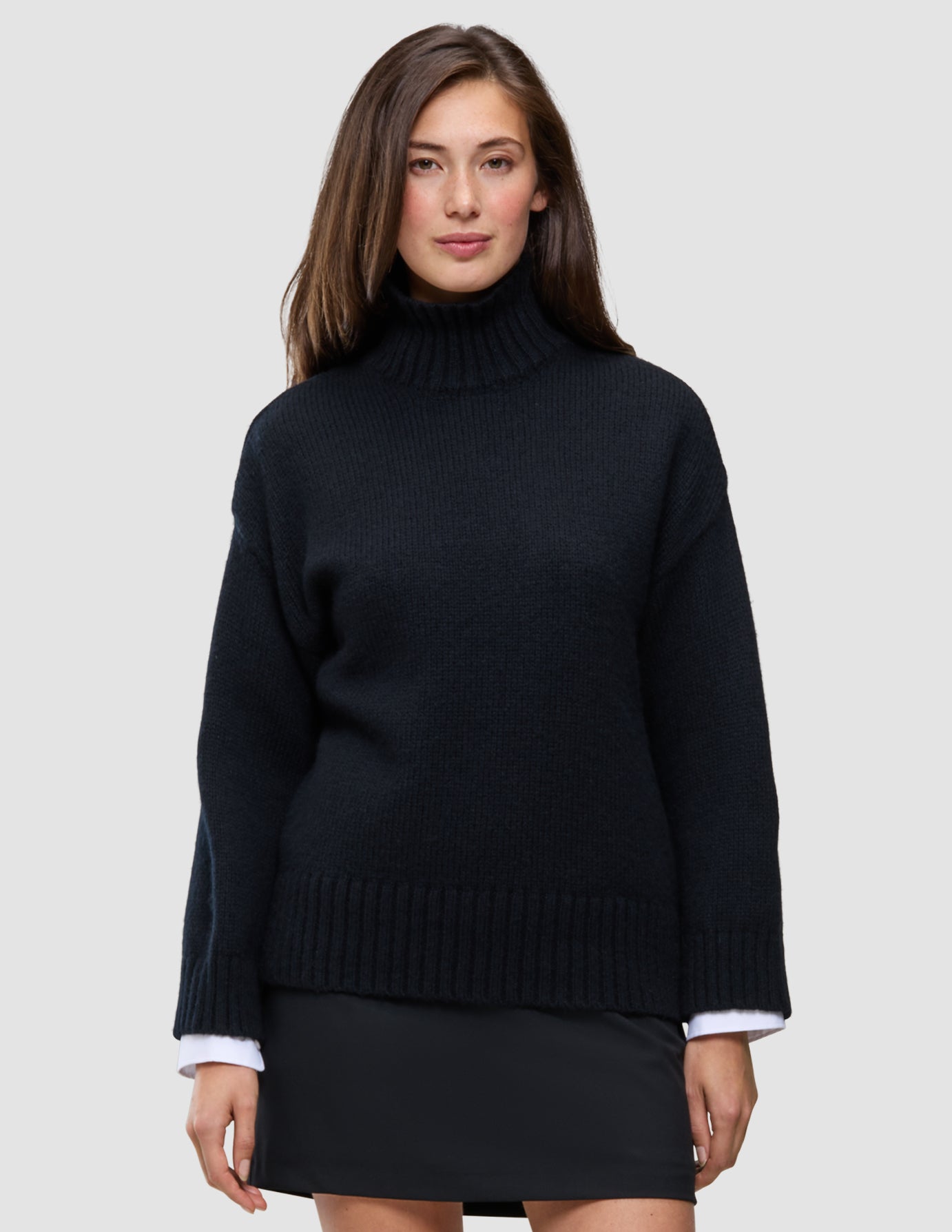 Eine Frau trägt den schwarzen, grob gestrickten, weichen Woll-Rollkragenpullover für Damen aus Merinowolle, an dessen Ärmeln weiße Hemdmanschetten hervorschauen.