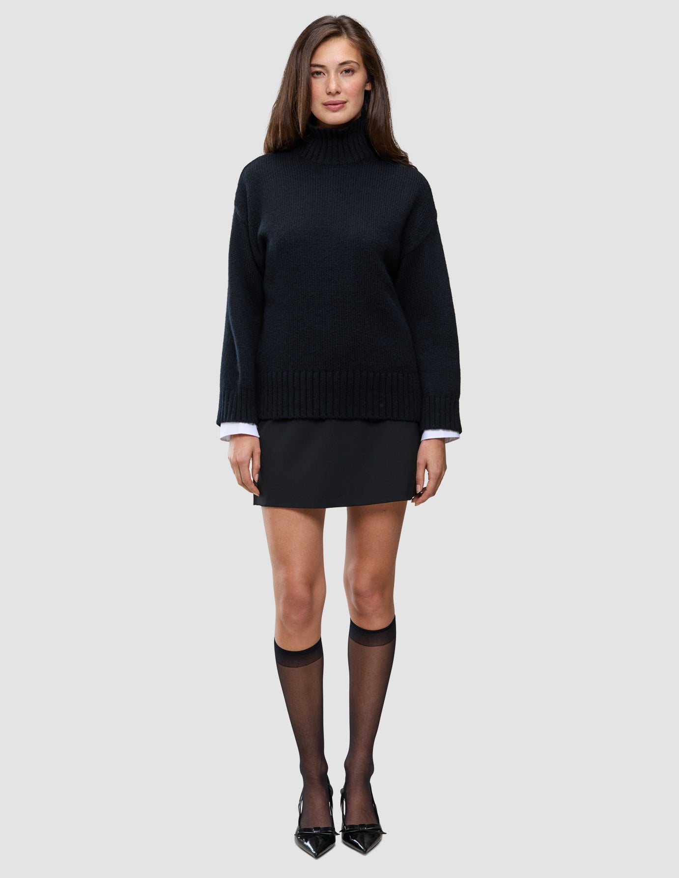 Eine Frau trägt einen schwarzen, grob gestrickten, weichen Wollpullover mit Rollkragen für Damen über einem weißen Hemd mit Kragen, dazu einen schwarzen Minirock, transparente schwarze Kniestrümpfe und schwarze Pumps.