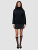 Eine Frau trägt einen schwarzen, grob gestrickten, weichen Wollpullover mit Rollkragen für Damen über einem weißen Hemd mit Kragen, dazu einen schwarzen Minirock, transparente schwarze Kniestrümpfe und schwarze Pumps.