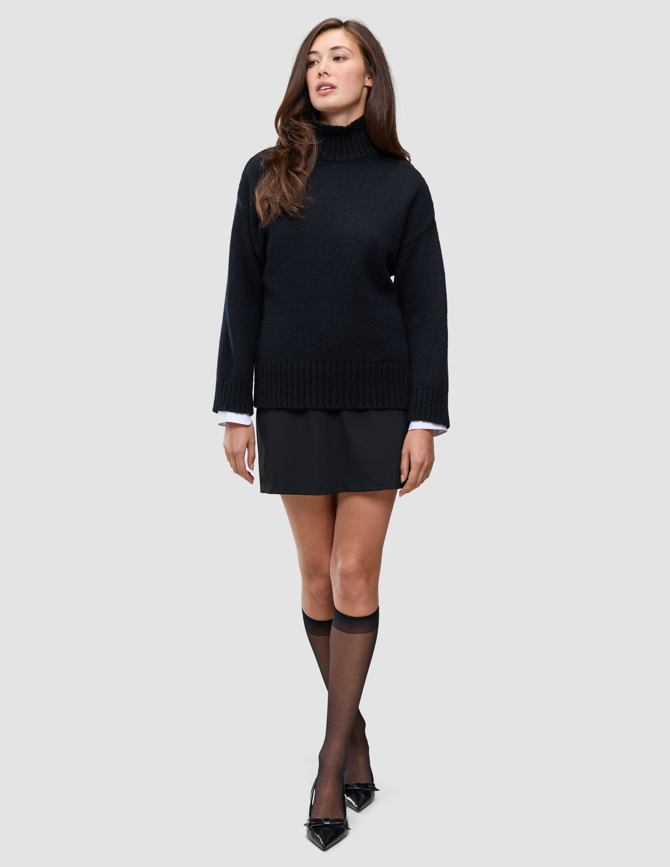 Eine Frau trägt einen schwarzen, grob gestrickten, weichen Wollpullover mit Rollkragen für Damen zusammen mit einem schwarzen Rock, schwarzen, durchsichtigen Kniestrümpfen und schwarzen Pumps.