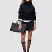Eine Frau, die einen schwarzen, grob gestrickten, weichen Wollpullover mit Rollkragen für Damen über einem weißen Hemd mit Kragen trägt, dazu einen schwarzen Minirock, transparente Kniestrümpfe, Kitten-Heels und eine schwarze Tragetasche.