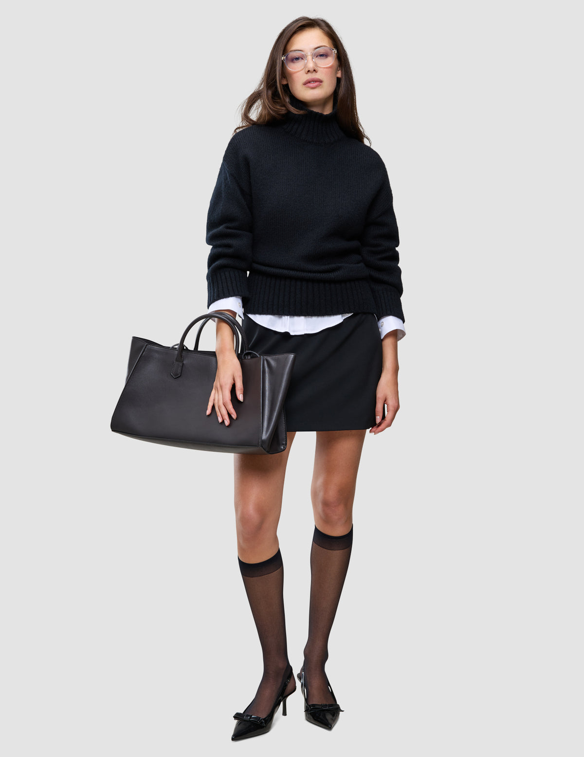 Eine Frau, die einen schwarzen, grob gestrickten, weichen Wollpullover mit Rollkragen für Damen über einem weißen Hemd mit Kragen trägt, dazu einen schwarzen Minirock, transparente Kniestrümpfe, Kitten-Heels und eine schwarze Tragetasche.