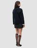 Rückansicht einer Person, die einen schwarzen, grob gestrickten, weichen Wollpullover mit Rollkragen für Damen, einen schwarzen Minirock sowie schwarze, transparente Kniestrümpfe und schwarze Pumps trägt.