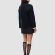 Rückansicht einer Person, die einen schwarzen, grob gestrickten, weichen Wollpullover mit Rollkragen für Damen, einen schwarzen Minirock sowie schwarze, transparente Kniestrümpfe und schwarze Pumps trägt.