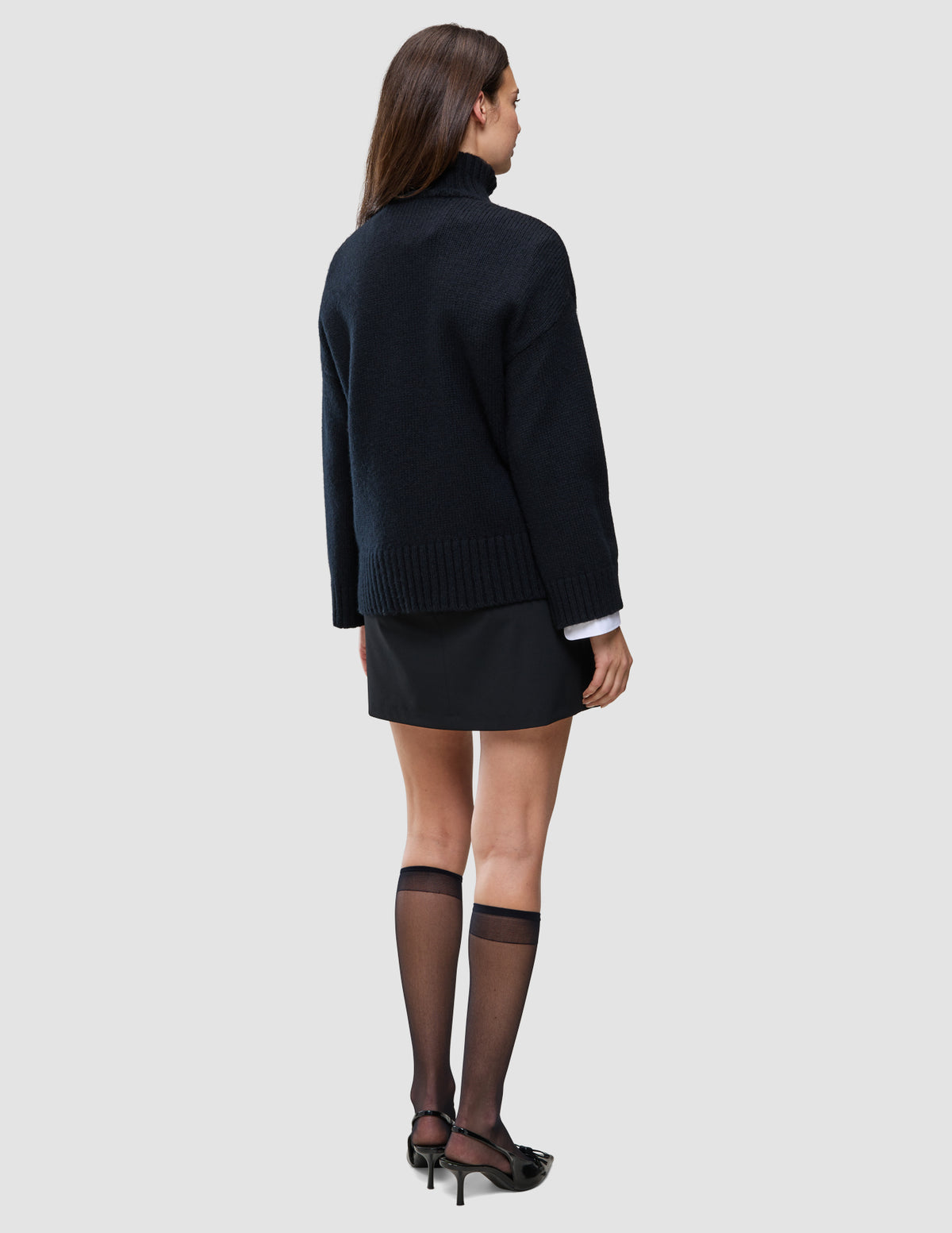 Rückansicht einer Person, die einen schwarzen, grob gestrickten, weichen Wollpullover mit Rollkragen für Damen, einen schwarzen Minirock sowie schwarze, transparente Kniestrümpfe und schwarze Pumps trägt.